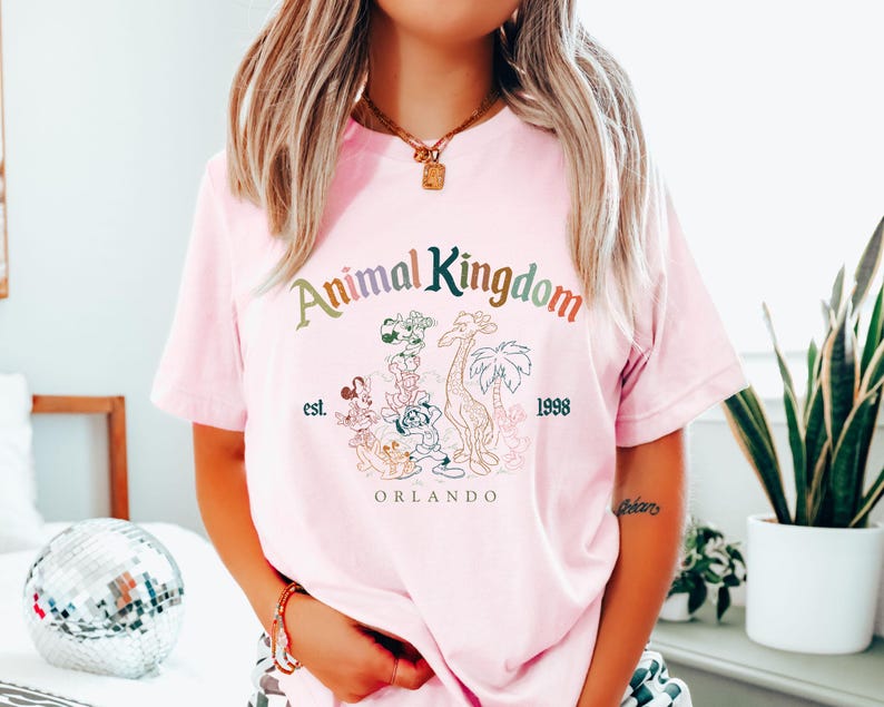 May include: A light pink t-shirt featuring the text "Animal Kingdom" in a multi-colored font, with a graphic of various cartoon animals. Below the text are the words "est. 1998" and "Orlando".