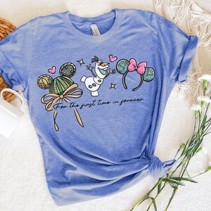 Puede incluir: Camiseta azul claro con un dise&ntilde;o de dibujos animados con orejas de Mickey Mouse, Olaf el mu&ntilde;eco de nieve y corazones. El texto "For the first time in forever" est&aacute; impreso debajo. La camiseta es de material suave.
