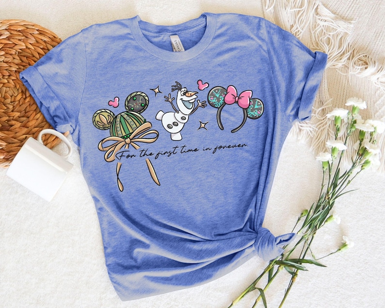 Puede incluir: Camiseta azul claro con un dise&ntilde;o de dibujos animados con orejas de Mickey Mouse, Olaf el mu&ntilde;eco de nieve y corazones. El texto "For the first time in forever" est&aacute; impreso debajo. La camiseta es de material suave.