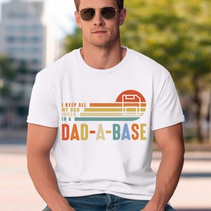 Könnte beinhalten: Weißes T-Shirt mit dem Text "I KEEP ALL MY DAD JOKES IN A DAD-A-BASE" in einem Retro-Design. Das Design verwendet ein Farbschema aus Rot, Orange, Gelb und Grün. Das T-Shirt hat einen Rundhalsausschnitt.