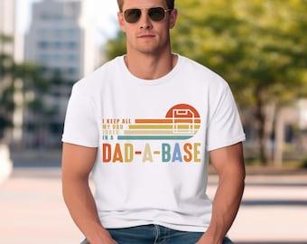 Op de afbeelding: Wit T-shirt met de tekst "I KEEP ALL MY DAD JOKES IN A DAD-A-BASE" in een retro-ontwerp. Het ontwerp gebruikt een kleurenschema van rood, oranje, geel en groen. Het T-shirt heeft een ronde hals.