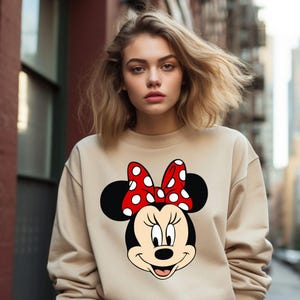 Puede incluir: Una sudadera beige con un lazo rojo con lunares blancos y una imagen de dibujos animados en blanco y negro de Minnie Mouse.