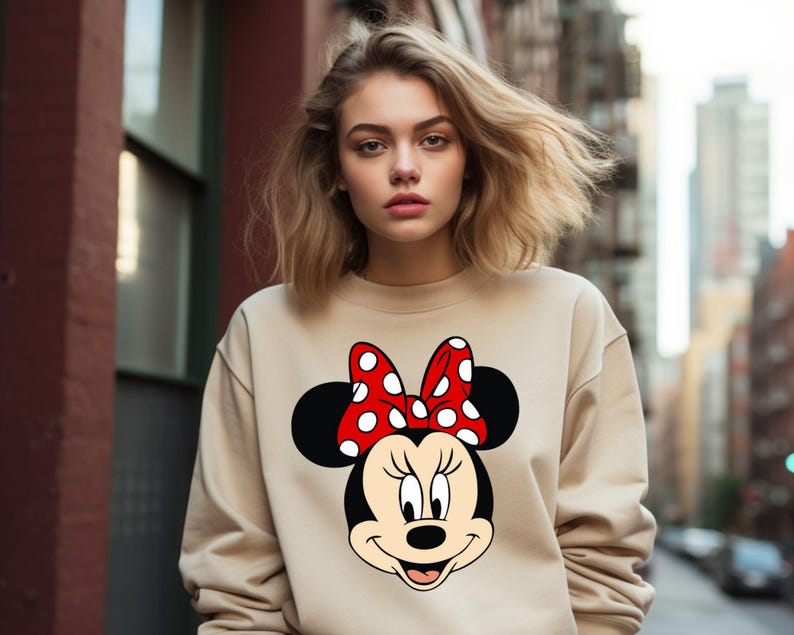 Puede incluir: Una sudadera beige con un lazo rojo con lunares blancos y una imagen de dibujos animados en blanco y negro de Minnie Mouse.