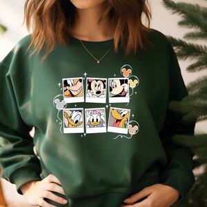 May include: A dark green sweatshirt with a graphic print of Mickey Mouse and friends in photo frames. The print includes Mickey Mouse, Minnie Mouse, Donald Duck, Goofy, and Pluto.