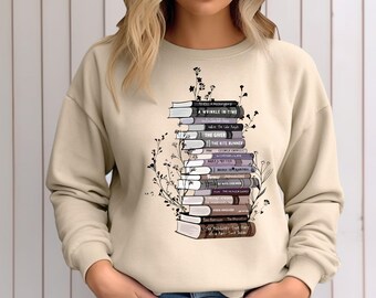 Puede incluir: Una sudadera beige con un estampado gráfico de una pila de libros con títulos como "Un pliegue en el tiempo", "El donante" y "El corredor de la cometa".