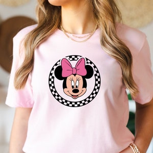 May include: Pink t-shirt with a black and white checkered circle featuring a cartoon image of Minnie Mouse with a pink bow.