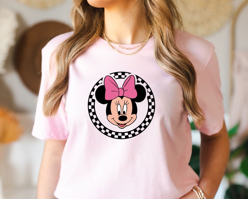 May include: Pink t-shirt with a black and white checkered circle featuring a cartoon image of Minnie Mouse with a pink bow.