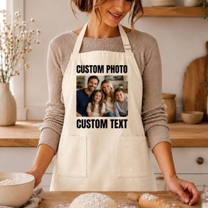 May include: A beige kitchen apron featuring a rectangular custom photo print of a family of four in the center. The text CUSTOM PHOTO is printed in black above the image, and the text CUSTOM TEXT is printed in black below the image. The apron has two front pockets.