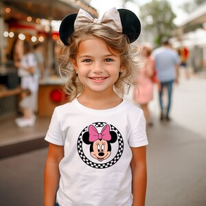 May include: A young girl wearing a white t-shirt with a black and white checkered circle featuring a cartoon image of Minnie Mouse with a pink bow. She is wearing black mouse ears with a gold bow.