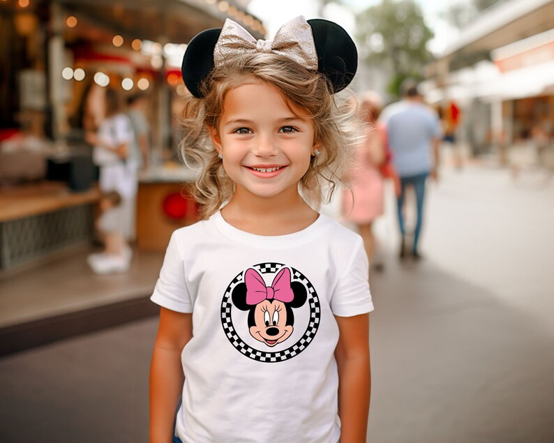 May include: A young girl wearing a white t-shirt with a black and white checkered circle featuring a cartoon image of Minnie Mouse with a pink bow. She is wearing black mouse ears with a gold bow.