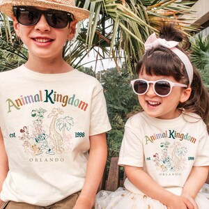 May include: Two children wearing cream-colored t-shirts with the words "Animal Kingdom" in rainbow colors. The shirts feature a graphic of cartoon animals and the text "Orlando" and "1998". The child on the left wears sunglasses and a straw hat.