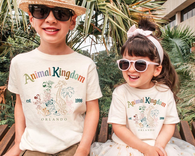 May include: Two children wearing cream-colored t-shirts with the words "Animal Kingdom" in rainbow colors. The shirts feature a graphic of cartoon animals and the text "Orlando" and "1998". The child on the left wears sunglasses and a straw hat.
