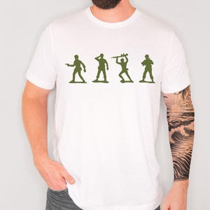 May include: White t-shirt with a green silhouette design of four toy soldiers in different poses. The soldiers are in a line. The shirt is made of a soft, comfortable material.