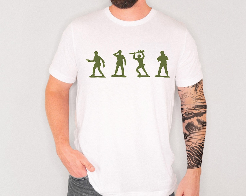 May include: White t-shirt with a green silhouette design of four toy soldiers in different poses. The soldiers are in a line. The shirt is made of a soft, comfortable material.