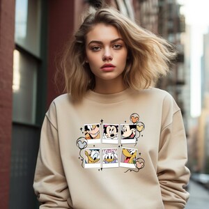 May include: A beige sweatshirt with a graphic print of Mickey Mouse and friends in a series of photo frames. The print includes Mickey Mouse, Minnie Mouse, Donald Duck, Goofy, and Pluto. The sweatshirt has a crew neck and long sleeves.