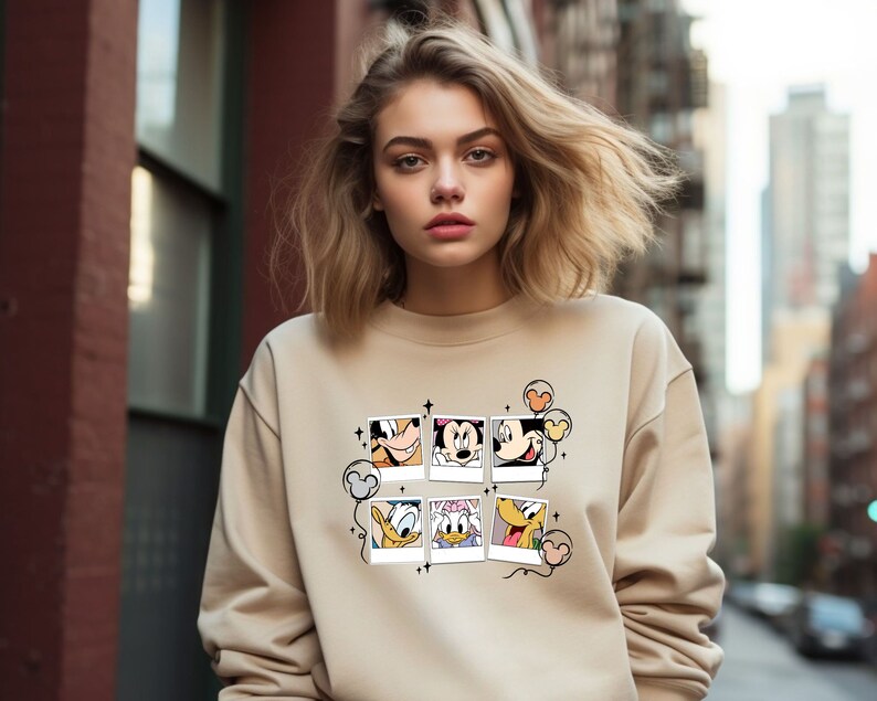 May include: A beige sweatshirt with a graphic print of Mickey Mouse and friends in a series of photo frames. The print includes Mickey Mouse, Minnie Mouse, Donald Duck, Goofy, and Pluto. The sweatshirt has a crew neck and long sleeves.