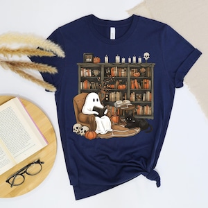 May include: Navy blue t-shirt with a Halloween illustration. A cartoon ghost reads a book in an armchair in front of a bookshelf. Pumpkins, candles, a skull, and a black cat are also featured. The shirt is designed for casual wear and comfort.