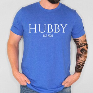 May include: A blue t-shirt with the word "HUBBY" in white capital letters, with "EST. 2025" below it. The shirt is made of a soft material and is being worn by a person.