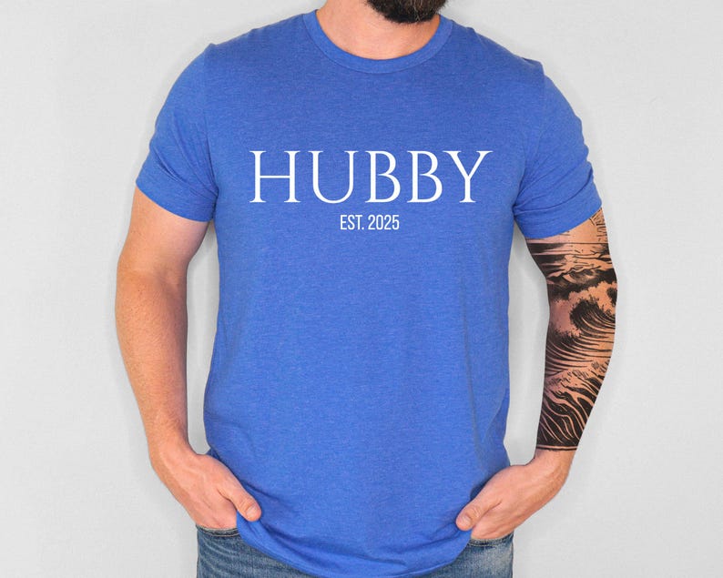 May include: A blue t-shirt with the word "HUBBY" in white capital letters, with "EST. 2025" below it. The shirt is made of a soft material and is being worn by a person.