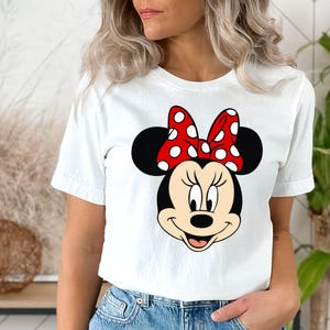 Puede incluir: Camiseta blanca con un lazo rojo con lunares blancos y una imagen de dibujos animados de la cara de Minnie Mouse.