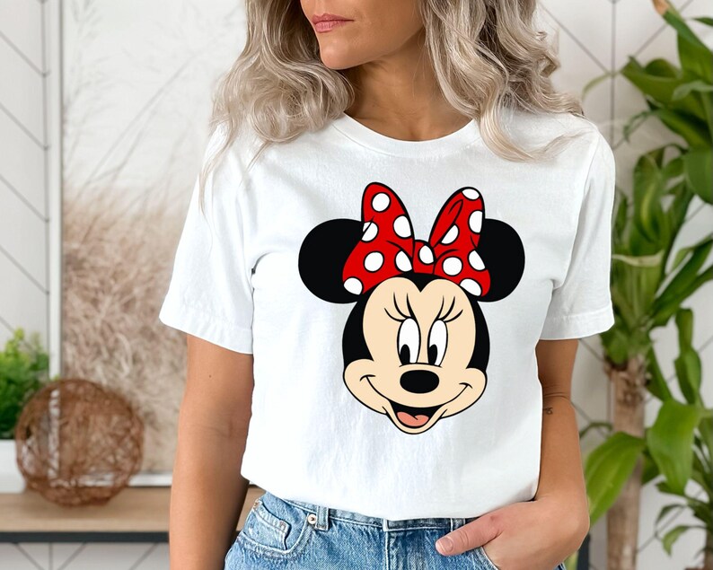 Puede incluir: Camiseta blanca con un lazo rojo con lunares blancos y una imagen de dibujos animados de la cara de Minnie Mouse.