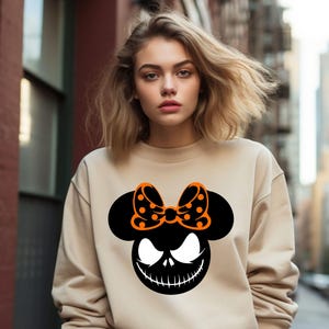 Puede incluir: Sudadera beige con un gr&aacute;fico negro y naranja. El gr&aacute;fico combina una silueta de Mickey Mouse con una cara de Jack Skellington y un lazo de lunares. Es una prenda informal de manga larga.