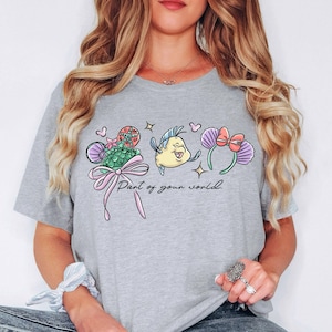 Puede incluir: Camiseta gris claro con un gr&aacute;fico colorido que presenta una cola de sirena, un pez amarillo y orejas de rat&oacute;n con un lazo rosa. El texto "Part of your world" est&aacute; debajo del gr&aacute;fico.