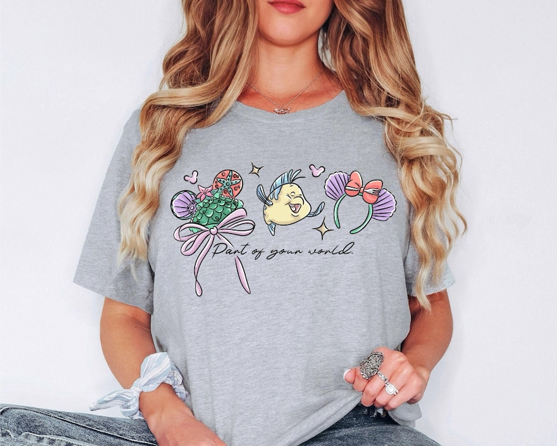 Puede incluir: Camiseta gris claro con un gr&aacute;fico colorido que presenta una cola de sirena, un pez amarillo y orejas de rat&oacute;n con un lazo rosa. El texto "Part of your world" est&aacute; debajo del gr&aacute;fico.