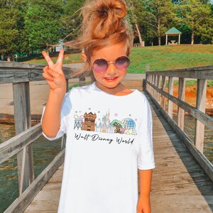 May include: White t-shirt with a colorful graphic of Walt Disney World landmarks, including a castle, Spaceship Earth, and other attractions. The words "Walt Disney World" are written in a script font below the graphic. The shirt is worn by a young person.