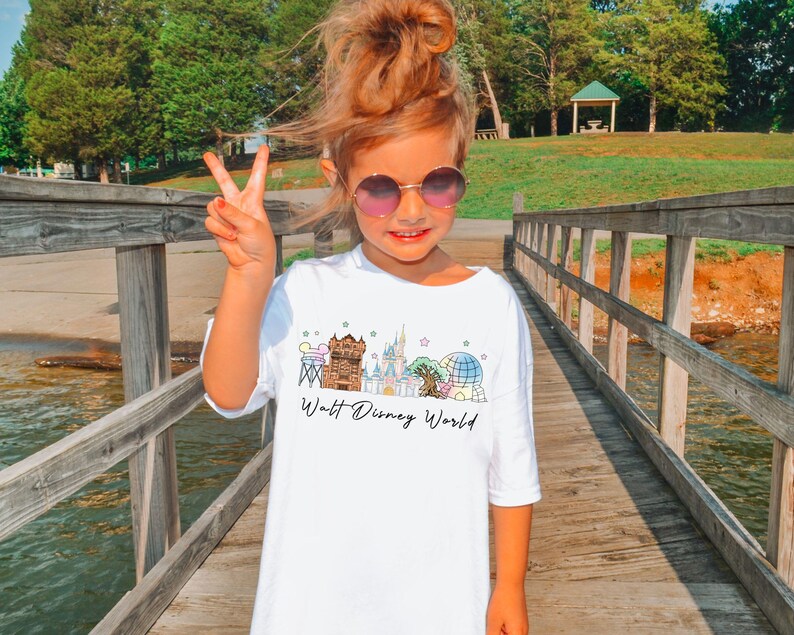 May include: White t-shirt with a colorful graphic of Walt Disney World landmarks, including a castle, Spaceship Earth, and other attractions. The words "Walt Disney World" are written in a script font below the graphic. The shirt is worn by a young person.