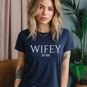 May include: Navy blue t-shirt with the word "WIFEY" in white, and the text "EST. 2025" below it. The shirt is a short-sleeved crew neck style.
