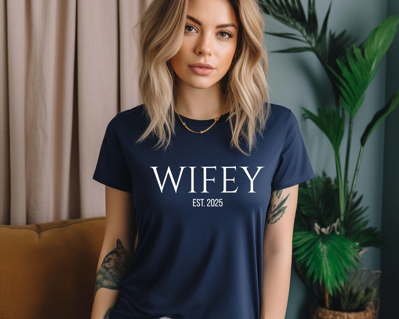 May include: Navy blue t-shirt with the word "WIFEY" in white, and the text "EST. 2025" below it. The shirt is a short-sleeved crew neck style.