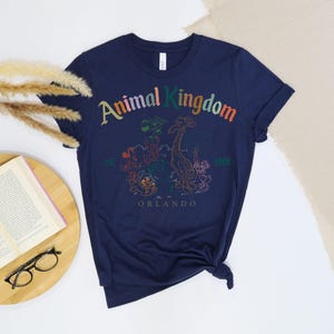 May include: Navy blue t-shirt with a colorful graphic of the Animal Kingdom logo and various animal illustrations. The text "Animal Kingdom" is arched across the top, with "est. 1998 Orlando" below the graphic. The shirt is knotted at the bottom.