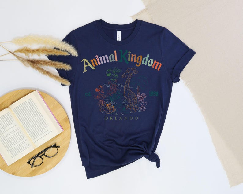 May include: Navy blue t-shirt with a colorful graphic of the Animal Kingdom logo and various animal illustrations. The text "Animal Kingdom" is arched across the top, with "est. 1998 Orlando" below the graphic. The shirt is knotted at the bottom.