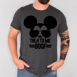 May include: A dark heather gray short-sleeve t-shirt featuring a black graphic of a mouse silhouette wearing sunglasses. Below the graphic, the text reads "TALK TO ME GOOF" in bold black capital letters, flanked by two horizontal black lines.