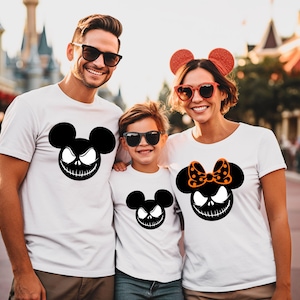Puede incluir: Tres personas vestidas con camisetas blancas con un dise&ntilde;o negro inspirado en Jack Skellington. El dise&ntilde;o presenta una forma de cabeza de Mickey Mouse con una sonrisa siniestra. La camisa de la mujer tiene un lazo naranja con lunares. La familia est&aacute; sonriendo.