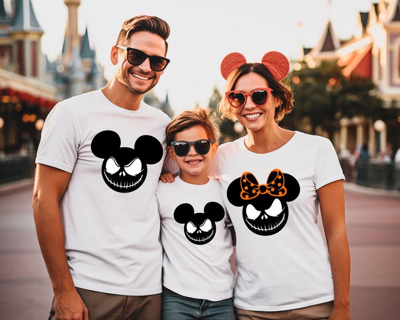 Puede incluir: Tres personas vestidas con camisetas blancas con un dise&ntilde;o negro inspirado en Jack Skellington. El dise&ntilde;o presenta una forma de cabeza de Mickey Mouse con una sonrisa siniestra. La camisa de la mujer tiene un lazo naranja con lunares. La familia est&aacute; sonriendo.