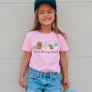 May include: A pink t-shirt featuring a colorful illustration of the Walt Disney World skyline. The design includes iconic landmarks and the text "Walt Disney World" in a script font. The shirt is worn by a young person.