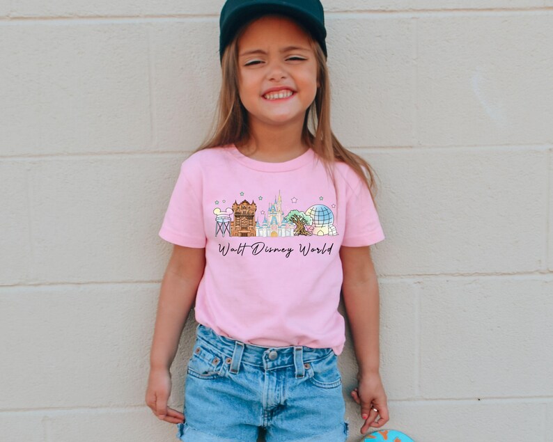 May include: A pink t-shirt featuring a colorful illustration of the Walt Disney World skyline. The design includes iconic landmarks and the text "Walt Disney World" in a script font. The shirt is worn by a young person.