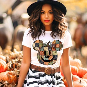 以下が含まれることがあります： ミッキーマウスの耳が付いたパッチワークのかぼちゃのデザインの白いTシャツ。 かぼちゃは笑顔で、カボチャに囲まれています。 その人は黒い帽子、茶色のベルト、白と黒のストライプのスカートを着ています。