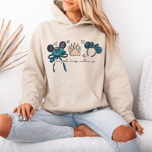 Puede incluir: Sudadera con capucha beige con un gr&aacute;fico frontal que presenta orejas de Mickey Mouse, una huella de pata y un lazo. El texto "Our fate lives within us" est&aacute; impreso debajo de los gr&aacute;ficos. La modelo lleva vaqueros azul claro.