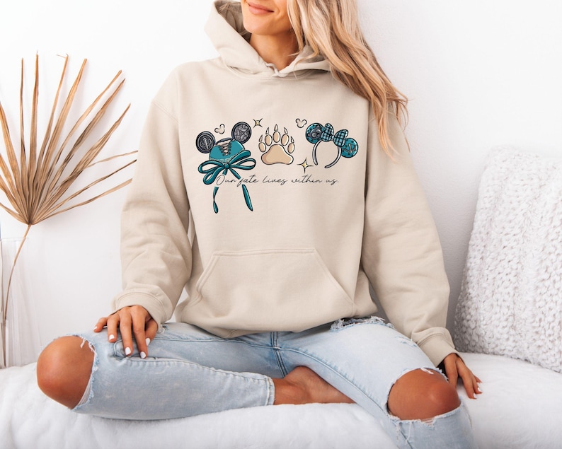 Puede incluir: Sudadera con capucha beige con un gr&aacute;fico frontal que presenta orejas de Mickey Mouse, una huella de pata y un lazo. El texto "Our fate lives within us" est&aacute; impreso debajo de los gr&aacute;ficos. La modelo lleva vaqueros azul claro.