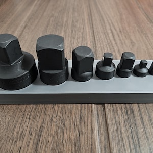 May include: A set of black, hexagonal impact sockets of varying sizes, arranged in a row on a gray tray. The sockets are made of metal and are designed for use with power tools. The tray is rectangular.