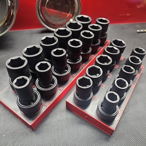Peut inclure: Ensemble de douilles à chocs noires disposées sur des plateaux rouges et gris. Les douilles, avec une ouverture hexagonale, sont conçues pour une clé. L'image présente différentes tailles, adaptées aux travaux automobiles ou mécaniques.