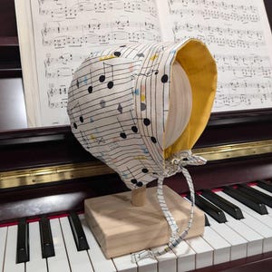 May include: A white and yellow bonnet with a musical note pattern. The bonnet is displayed on a wooden stand and a piano keyboard.