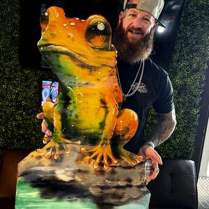 May include: A man with a beard and a grey baseball cap holds a large, colorful, wooden frog sculpture. The frog is sitting on a rock with water painted around it.