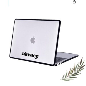 Könnte beinhalten: Ein schwarzes Laptop-Case mit einem weißen Laptop im Inneren. Der Laptop hat ein schwarzes Apple-Logo und das Wort "ainsley" ist auf dem Case aufgedruckt.