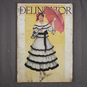 May include: Vintage magazine cover of "The Delineator" from 1910. The cover features a woman in a white dress with black trim, holding a pink parasol. The magazine title is in large, elegant lettering, with the date and issue details visible.
