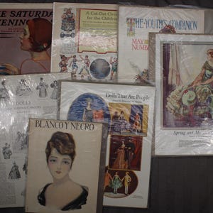 May include: A collection of vintage magazines and prints, including "The Saturday Evening," "The Youth's Companion," and "Blanco y Negro." The covers feature illustrations of women, children, and various scenes. The items are wrapped in plastic.
