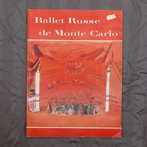 May include: A vintage book cover for "Ballet Russe de Monte Carlo" in red and white. The cover features an illustration of a stage with dancers and an audience, with the title at the top.
